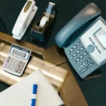 Meja kantor tersusun rapi dengan kalkulator, telepon, stapler, dan alat tulis, menggambarkan equipment atau peralatan kerja